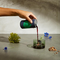 A person pouring a can of De Soi Purple Lune into a small cocktail glass with a blackberry garnish.