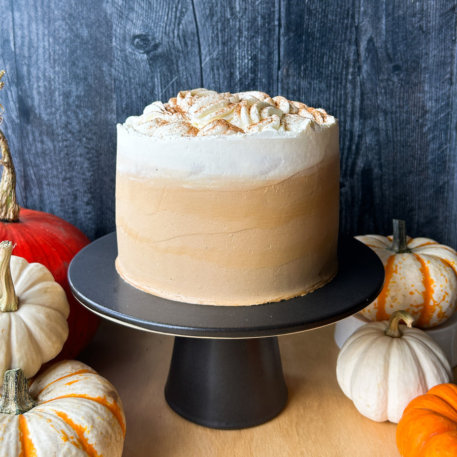 A cake frosted in white buttercream, with a diffuse band of light brown buttercream along the bottom. The cake is topped with whipped cream and a dusting of cinnamon.