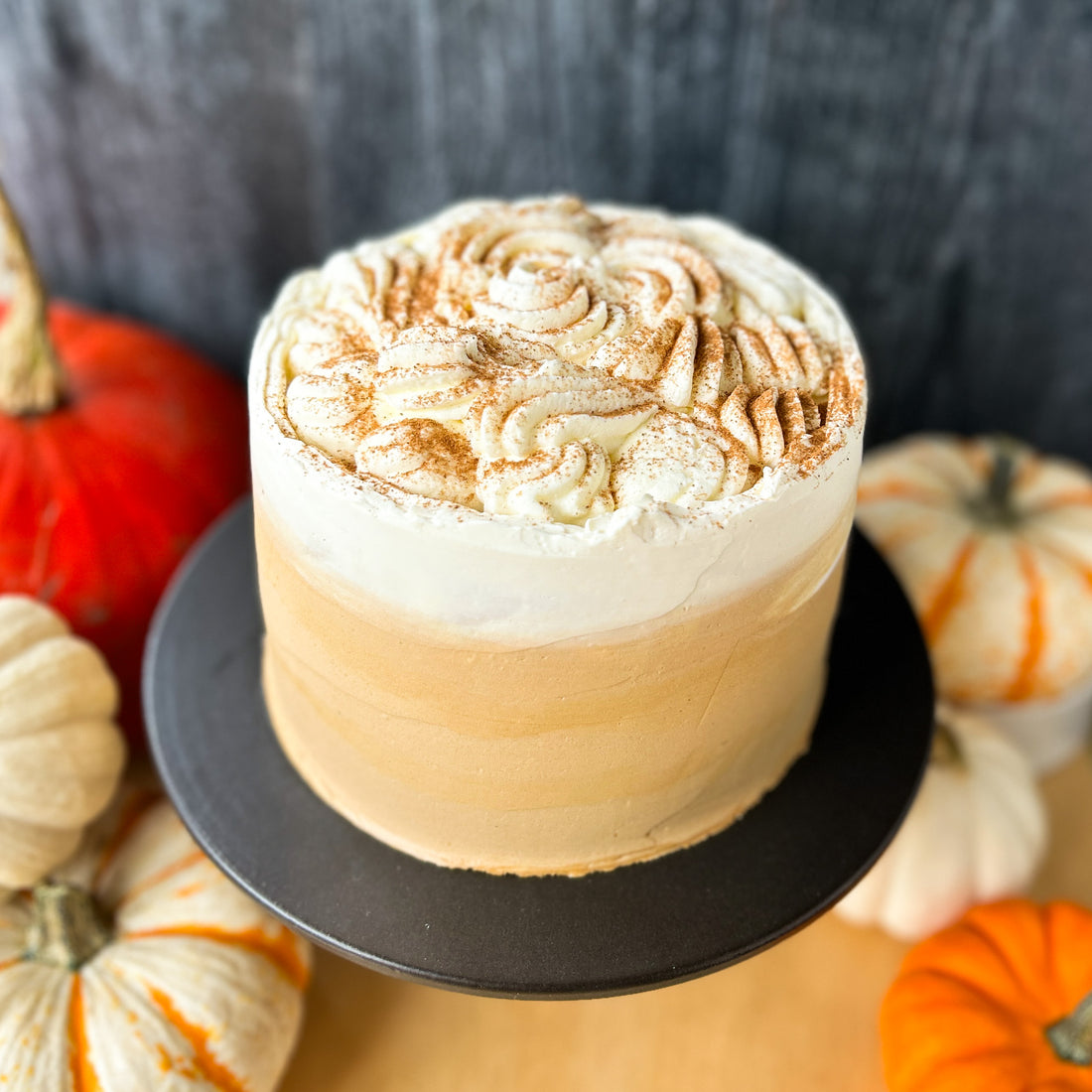 A cake frosted in white buttercream, with a diffuse band of light brown buttercream along the bottom. The cake is topped with whipped cream and a dusting of cinnamon.