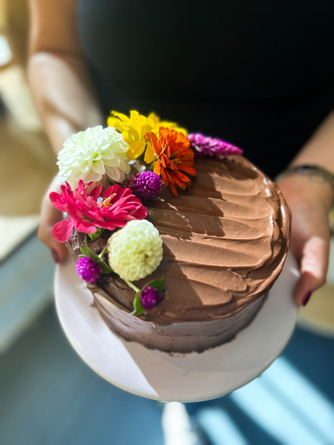 A  person holding a round cake frosted with fudgey buttercream and decorated with a crescent of fresh flowers.