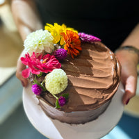 A  person holding a round cake frosted with fudgey buttercream and decorated with a crescent of fresh flowers.