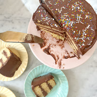 A round cake frosted with fudgey buttercream and decorated with colorful sprinkles. The cake is cut into, revealing layers of yellow cake and more chocolate buttercream.