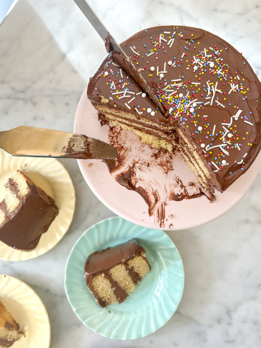 A round cake frosted with fudgey buttercream and decorated with colorful sprinkles. The cake is cut into, revealing layers of yellow cake and more chocolate buttercream.