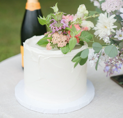 A tall round cake frosted with rustic white buttercream and and topped with a colorful floral bouquet.