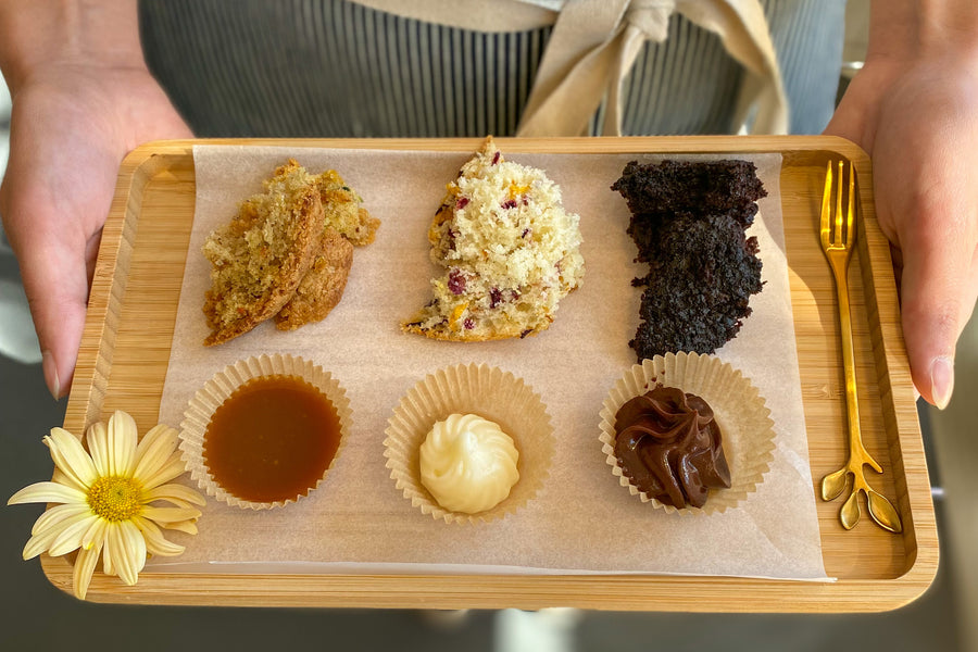 A person holding a board with three piles of cake scraps and three ramekins of frosting, all different flavors.